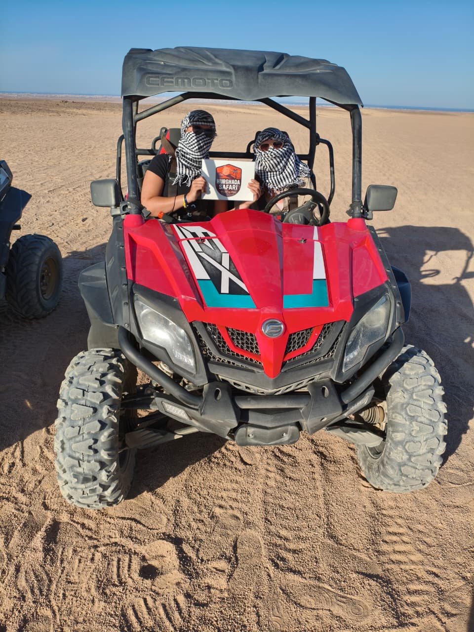 Hurghada Safari guests in a red CFMOTO buggy holding the Hurghada Safari sign in the desert